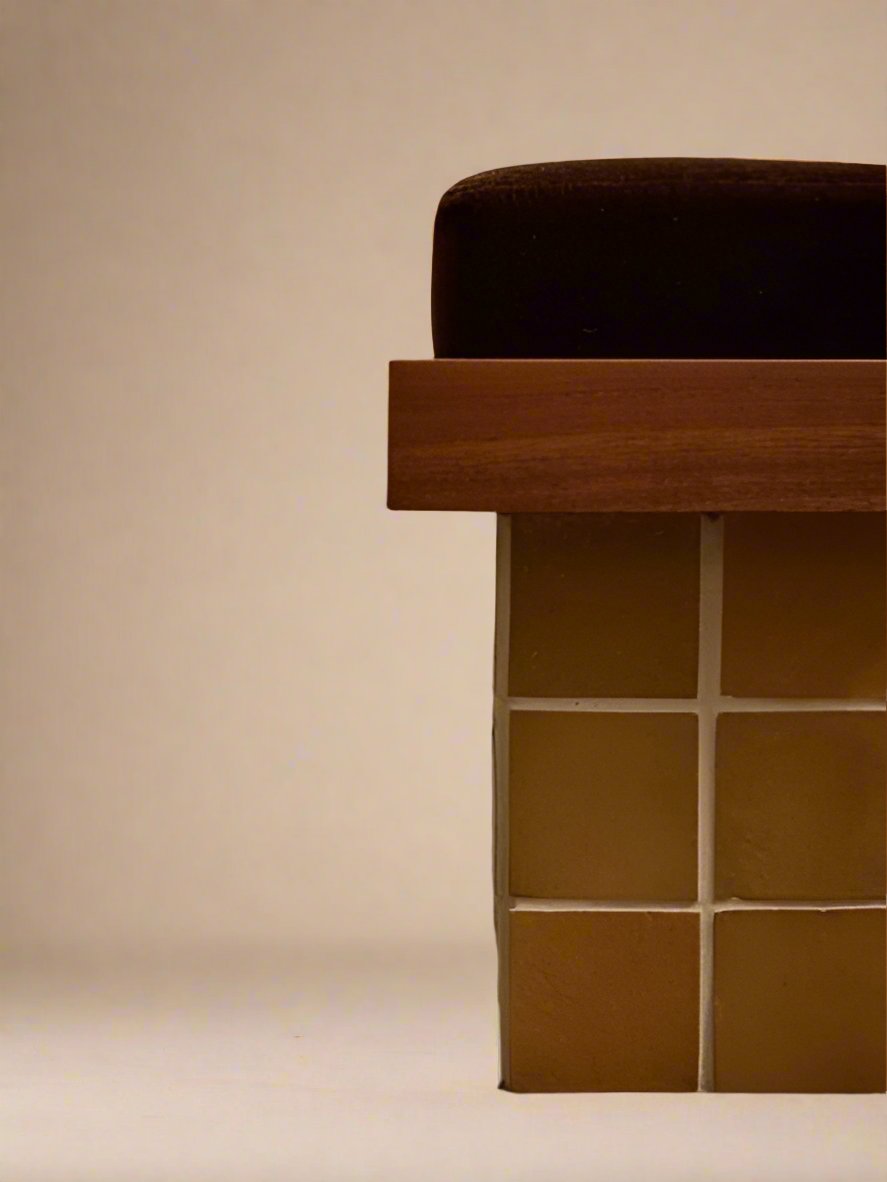 The Jura Stool by Ah Um Design Studio is shown in close-up, highlighting its brown cushion, wooden edge, and square yellow-tiled base set against a plain beige background.