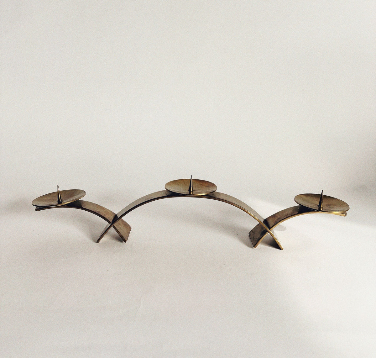 The Huerto 2 Curved Brass Candleholders feature a modern, arched design inspired by mid-century style, each with flat round plates for candles, shown against a plain white background.