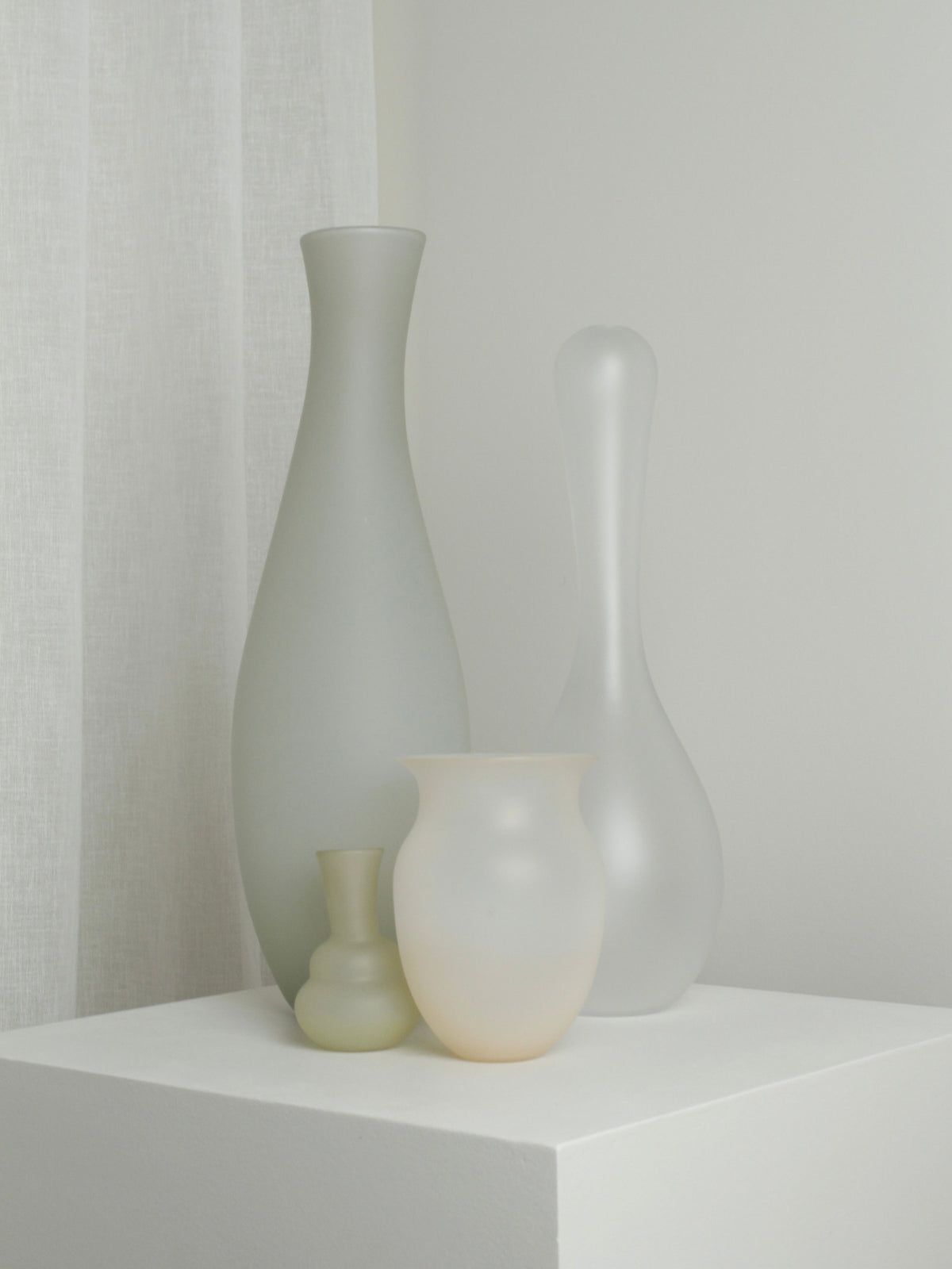 Four frosted glass vases, including Verre d'Onge's XLarge Vase in Ghost, are displayed on a white pedestal in a softly lit, minimal room with white walls and a sheer curtain.