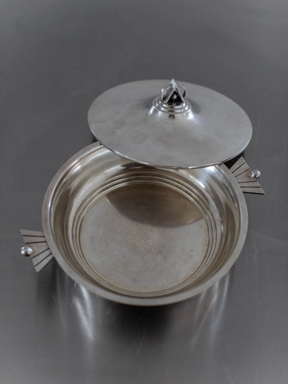 The Out For Lunch Art Deco Bowl, a round silver piece with geometric side handles and a partially open lid, exudes vintage glamour as it rests on a reflective metallic surface.