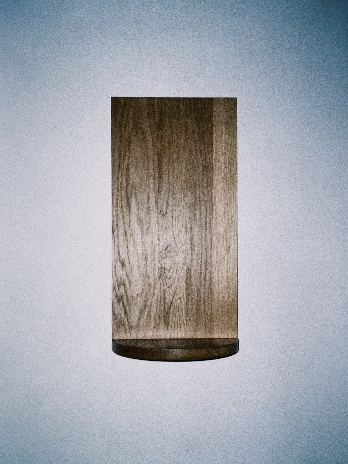 The Megaron - Minimalist Shelf in Wood - Small by COSEINCORSO is a rectangular handcrafted solid oak shelf with a curved base and visible grain, mounted on a light blue wall with softly lit, gradient background.