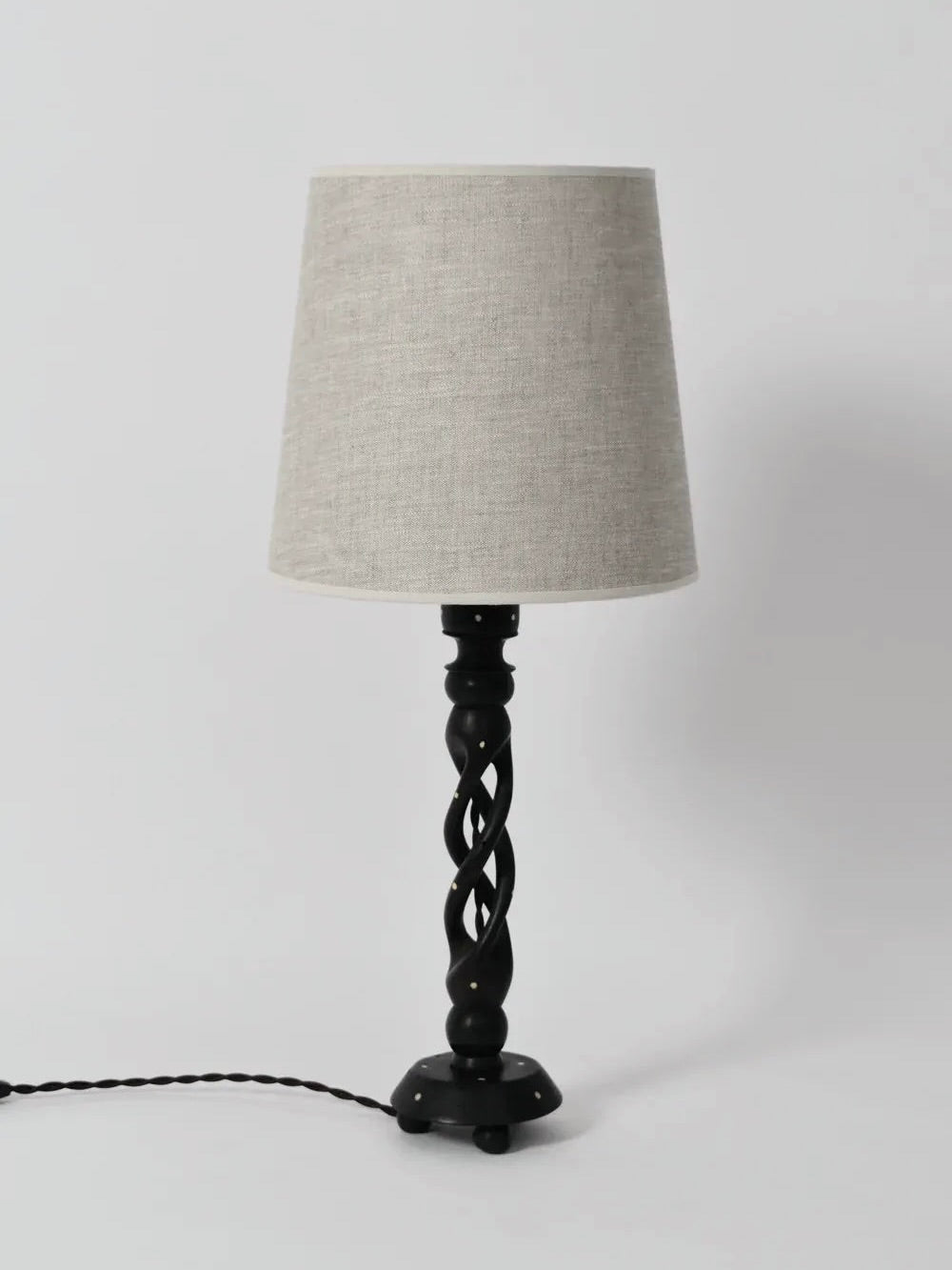 The Turned Wood Twist Lamp by Barracuda Interiors (1960s) features a black twisted base and light beige fabric shade, standing on a white surface with its black power cord extending to the left.