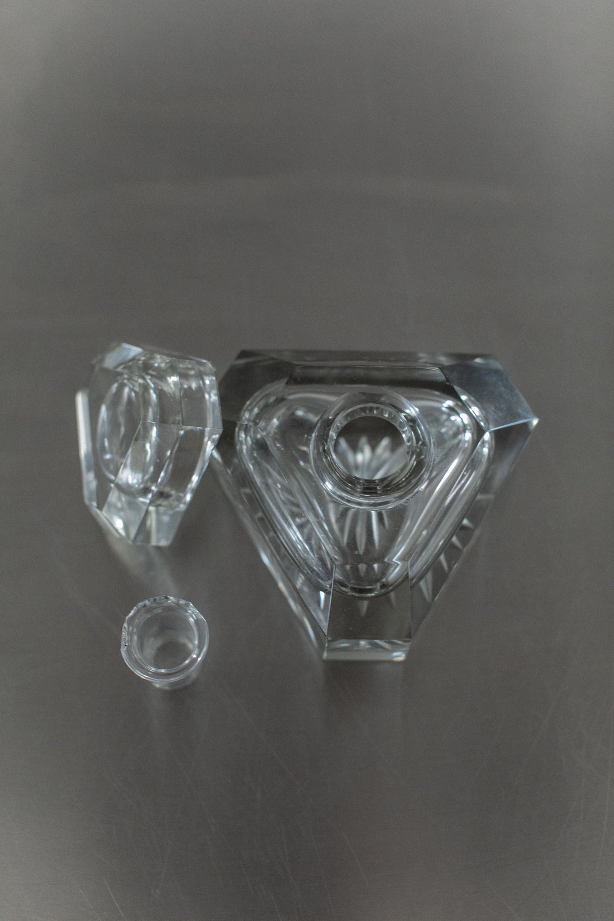 A geometric, diamond-shaped Glass Perfume Bottle by Out For Lunch, with exquisite craftsmanship, sits on a gray surface. Its matching glass lid has been removed and placed nearby, alongside a small round glass insert.