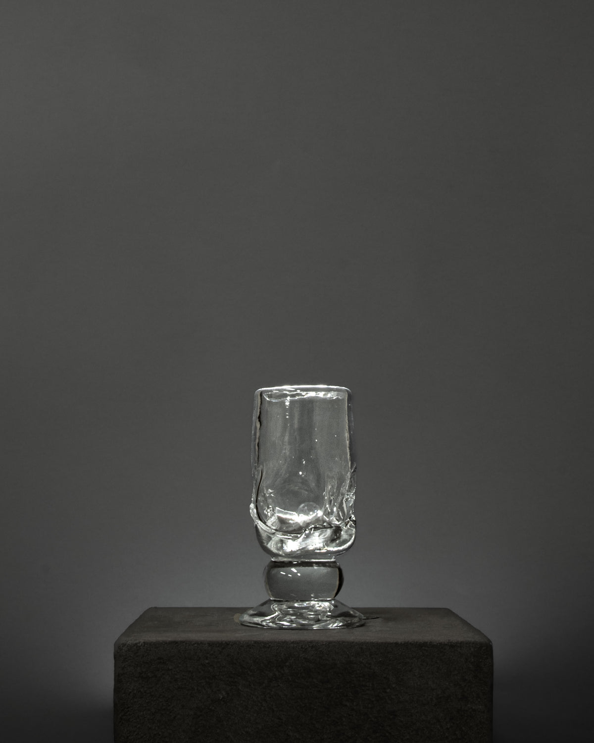 An empty Organic Footed Table Glass by Szkło Studio, hand-blown with a textured surface, stands on a dark stone pedestal against a plain, dark gray background.