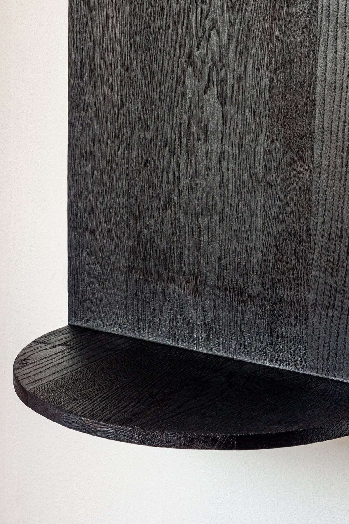 A close-up of COSEINCORSO’s The Megaron - Minimalist Shelf in Wood (Natural Oak Large) showcases its dark, circular form and visible oak grain, handcrafted and mounted on a matching vertical panel against a white background.