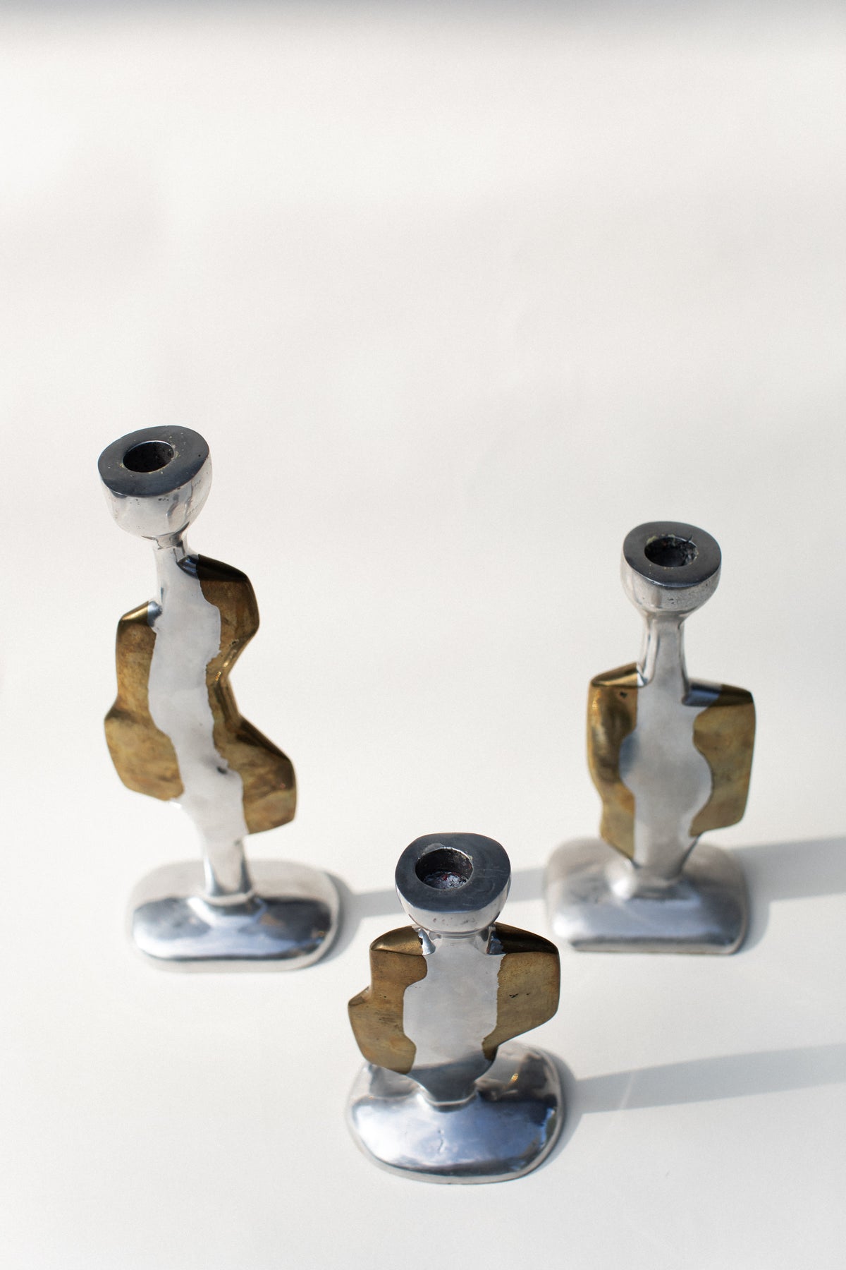 The Three Brutalist Candleholders by Out For Lunch are modern, abstract aluminum holders in silver and brass tones with geometric, sculptural shapes and varying heights, arranged on a light surface casting soft shadows.
