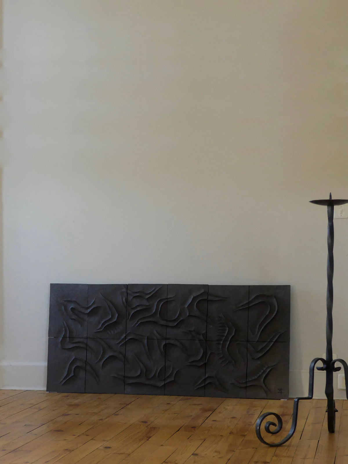 The Caldeira Wall Sculpture by Franck Scala, a dark abstract ceramic bas-relief, leans against a beige wall on a light wooden floor. Nearby stands a tall, slender black metal stand with a circular top.