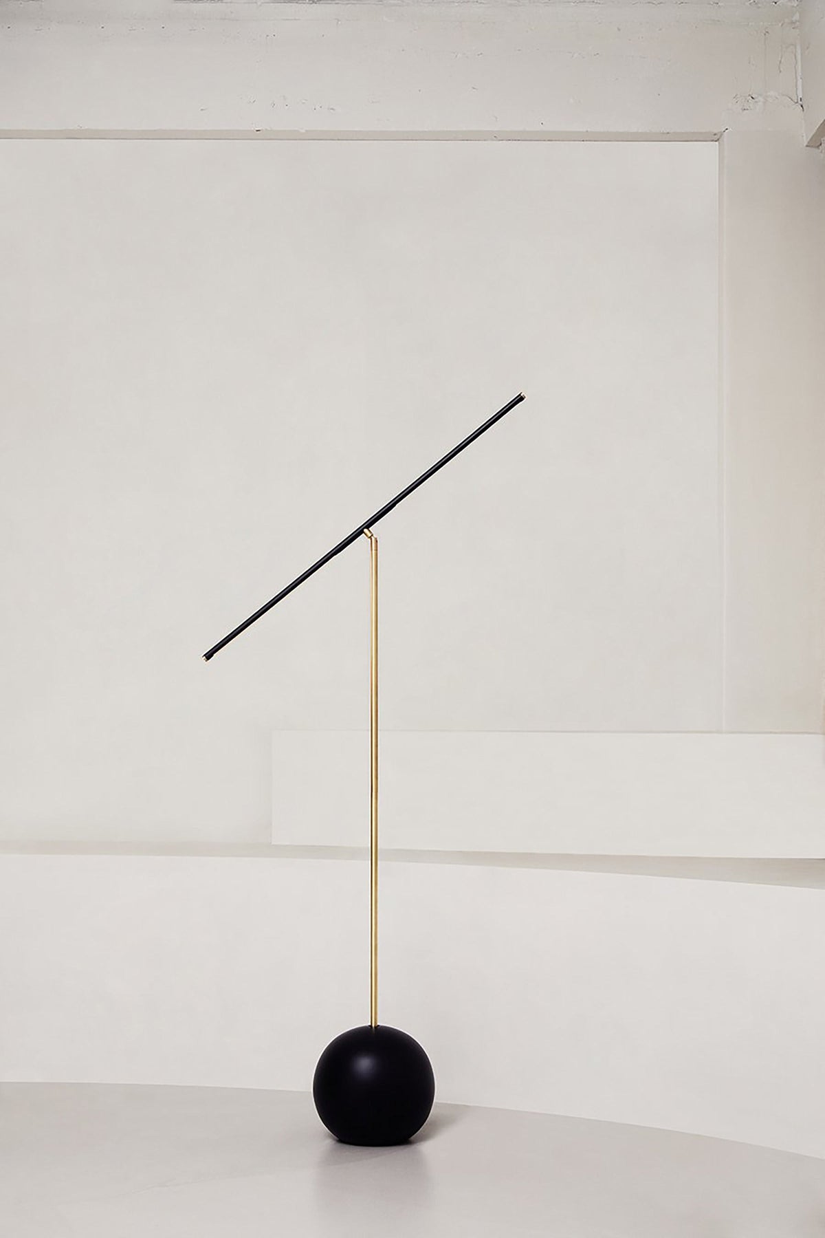 The 'T-Lamp' Floor Lamp by Heako Studio features a thin gold pole, round black base, and a long diagonal black light bar for striking sculptural lighting against a white wall.