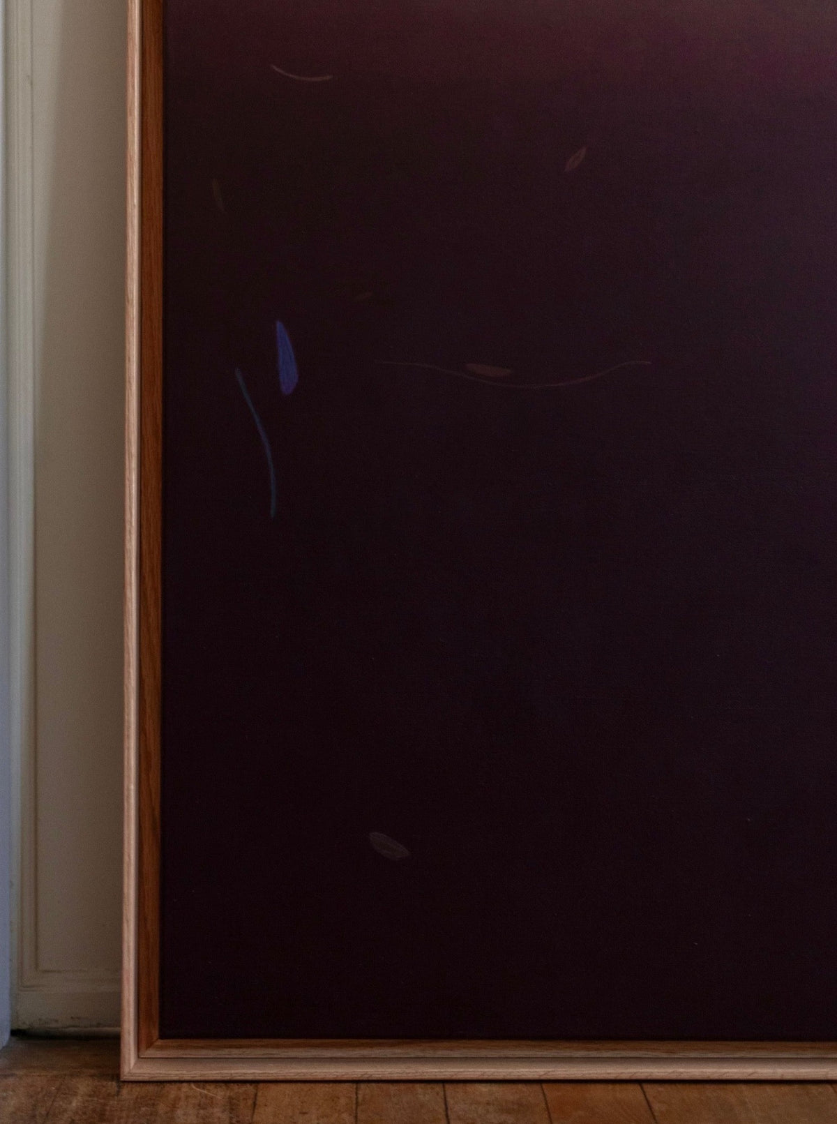 A painting with a hard waxed oak frame leans against a white wall. The artwork features a gradient of dark hues, primarily purples and browns, with subtle abstract shapes and lines. Acrylics on canvas, "Something(s) in the air, 2024" by Rebecca von Matérn rests on a wooden floor.