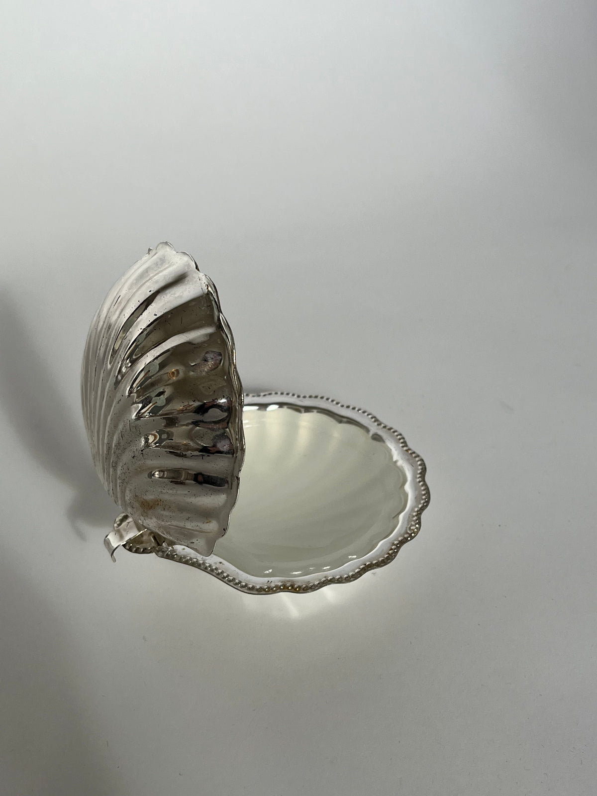 The septembre studios Shell Shaped Butter Dish, featuring a silver finish, hinged lid, and decorative textured edge, is shown partially open against a plain white background for an elegant display.