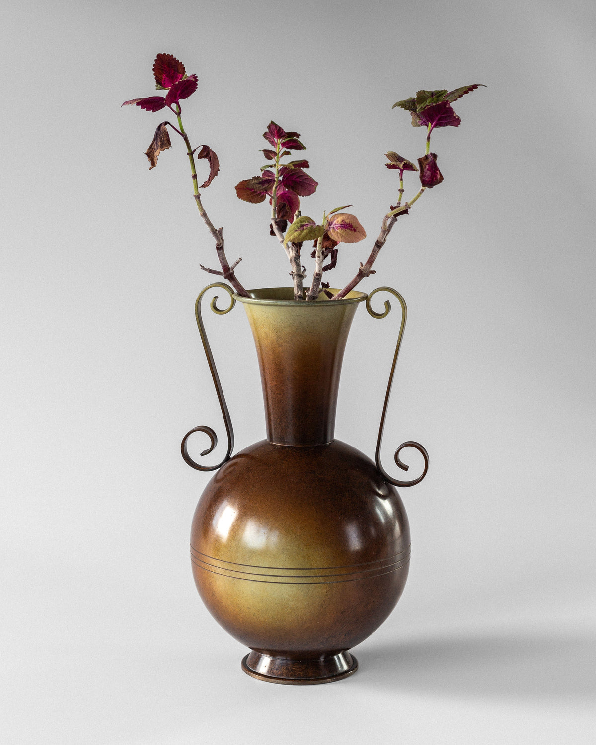 The Spigel Swedish Art Deco Bronze Vase features a brown, round-bodied design with elegant curved handles and holds branches with deep red and green leaves, set against a plain light gray background.