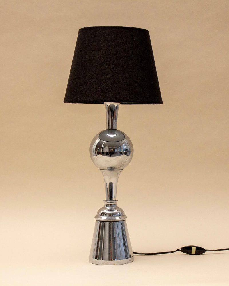 The SÒLER ALBUFEIRA BLACK LAMP features a shiny metal base with a round center detail and a black lampshade. The lamp, switched off, stands against a plain beige background.