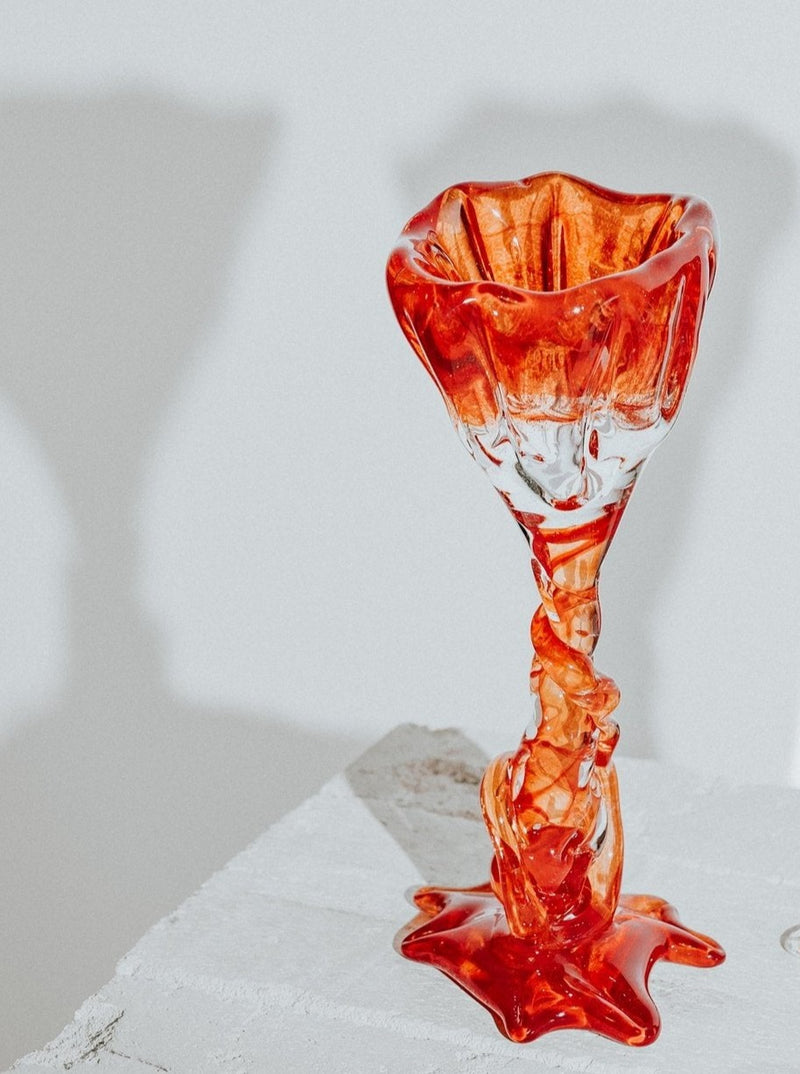 A sculptural Spiky Cocktail Glass - Red #1 by Szkło Studio with a twisted stem, melting onto a white surface, creating a vivid, liquid-like pool at its base. The background is softly focused in neutral tones.
