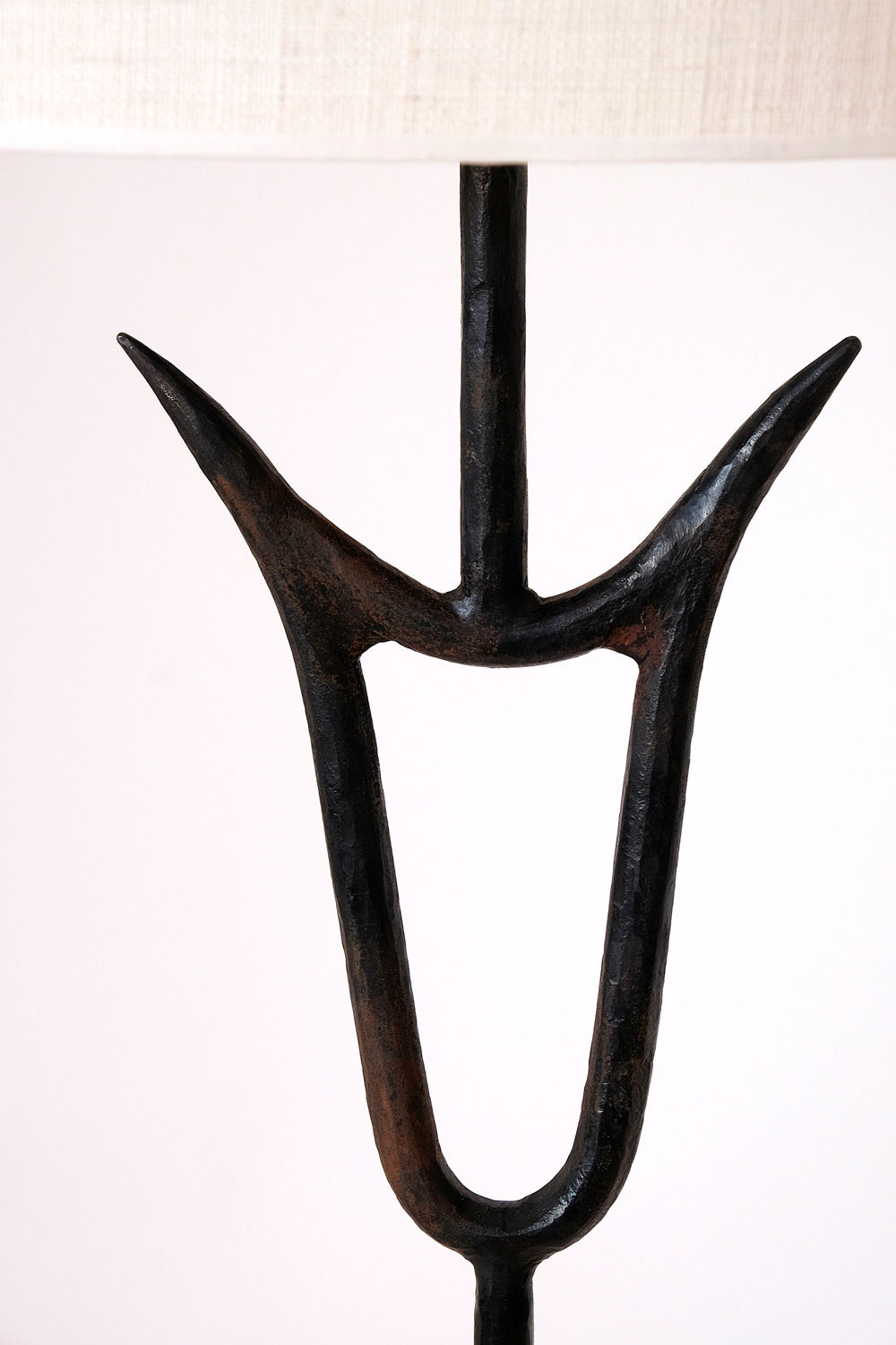 A close-up of the "Sevilla" Lamp by Barracuda Interiors shows its zoomorphic hammered iron base with a dark, textured finish, featuring an abstract symmetrical design of upward-curving prongs and a central vertical support against a plain background.