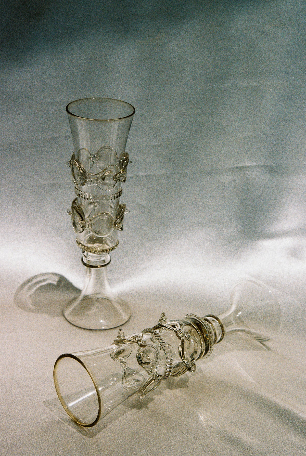 Two Lucie Claudia Podrabska mouthblown champagne flutes from the Dots Spirals collection, featuring gold rims and swirling details; one stands upright while the other rests on its side on a silky, reflective surface.