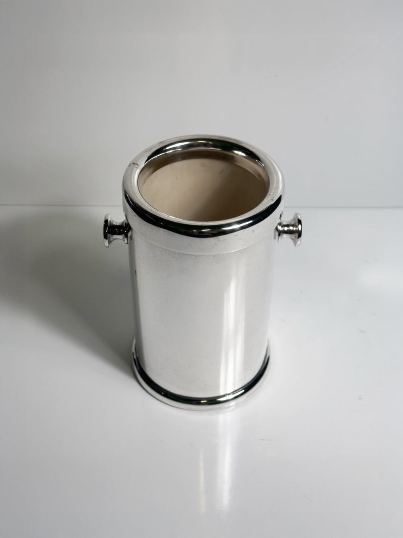 The Dodo Vintage Silver Plated Champagne Cooler - 1980s features a shiny, cylindrical design with two small side handles. Shown empty, it sits on a white surface against a plain white background.