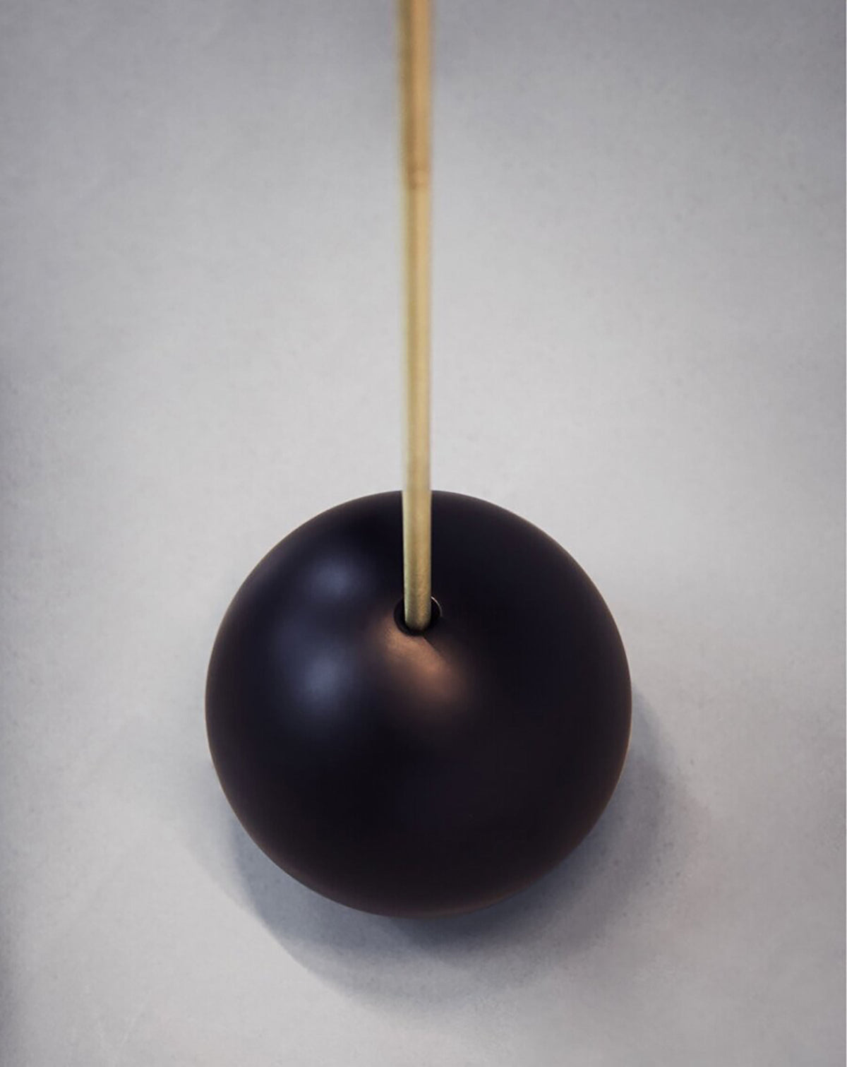 The 'T-Lamp' Floor Lamp by Heako Studio features a dark, glossy spherical base with a smooth finish and a slender vertical wooden stick, resembling a sculptural light, set against a light grey background.