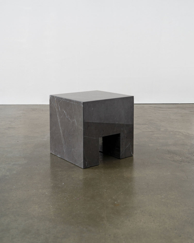 The Tom Bogle Postmodern Solid Marble Stool / Side Table is a small, rectangular dark stone piece with a geometric cutout at the base, giving it a sculptural look. It rests on a polished concrete floor against a plain light background.