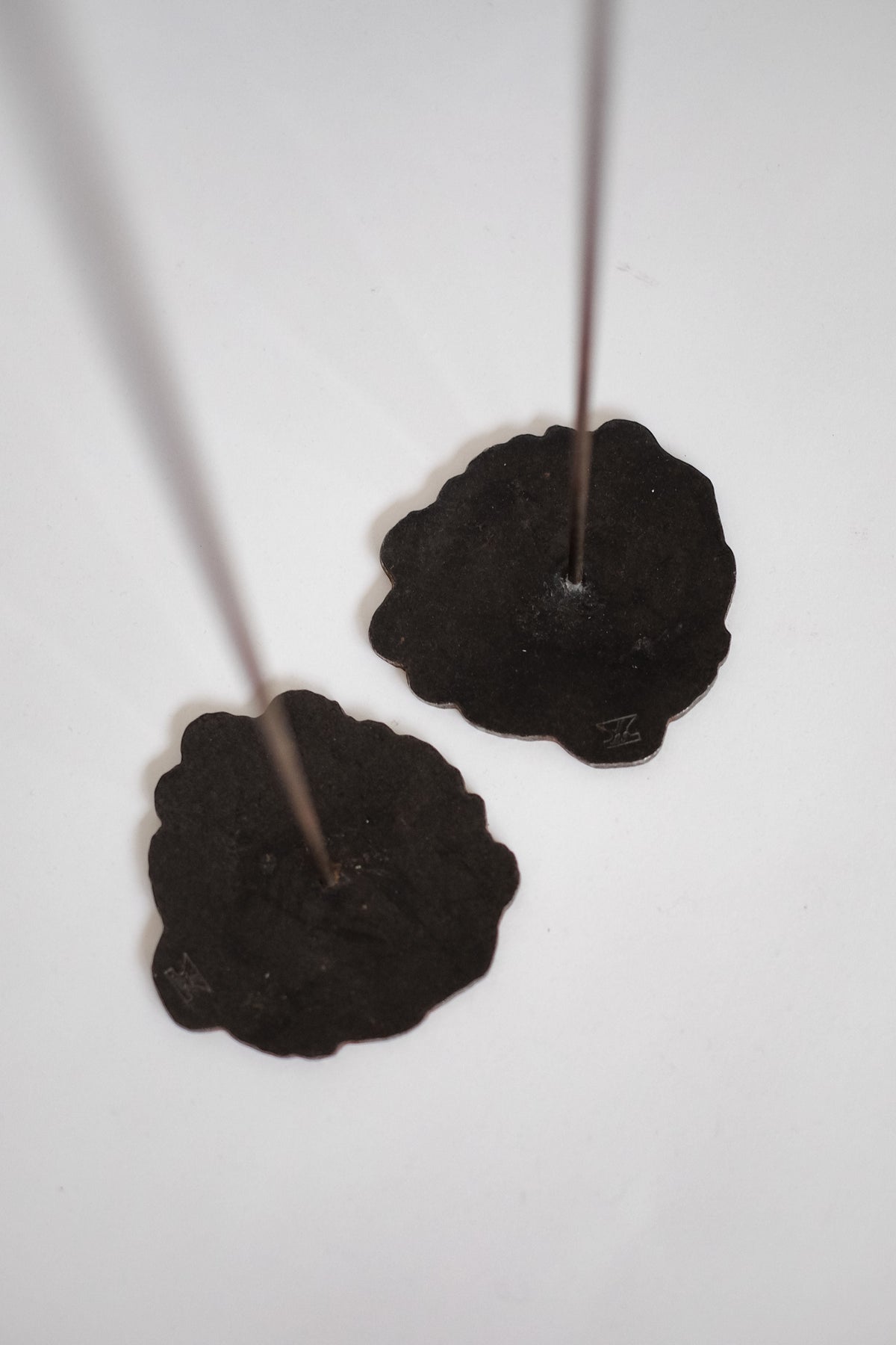 Two slim candles stand upright in Pieter Spruyt Copper Candle Scones by septembre studios, with flat, irregular black holders inspired by 1980s handmade designs, set against a plain white background.