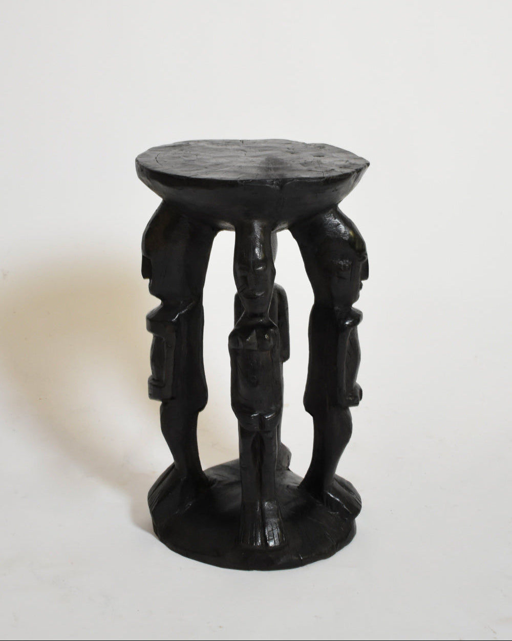 The African Anthropomorphic Side Table by Barracuda Interiors (1960s) features three carved human figures as legs supporting a round, uneven seat, crafted from dark ebonised wood and photographed against a white background.