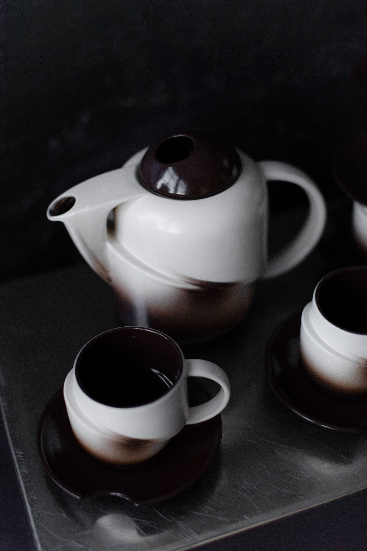 The Out For Lunch Art Deco Ceramic set—a teapot with matching teacup and saucer in a white-to-brown gradient—exudes vintage serveware elegance on a reflective metal surface against a dark background.