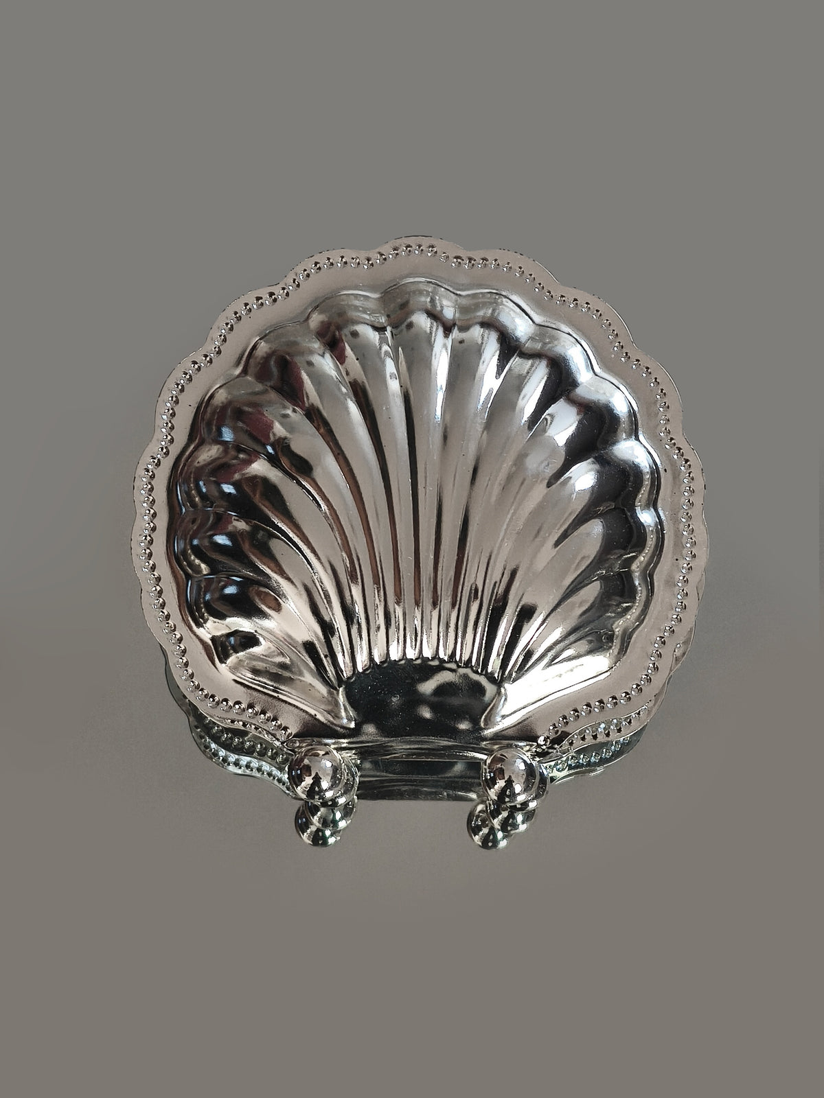 The Les Objoies Napkin Shell Holder features a silver-plated, scallop shell design with a beaded edge and three rounded feet—perfect for bringing seaside charm to your table. Displayed on a plain gray background.