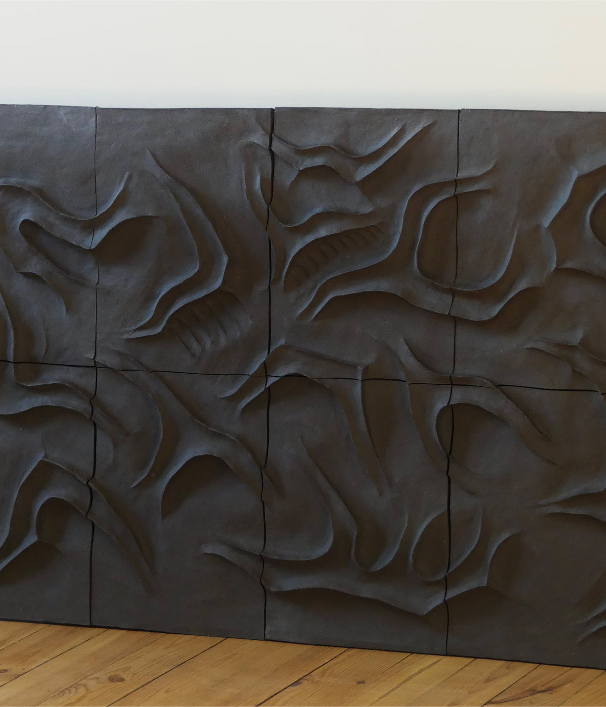 The Caldeira Wall Sculpture by Franck Scala is a dark, sculpted ceramic panel with an abstract, organic relief design of swirling raised shapes and grooves, displayed against a light wall above a wooden floor.