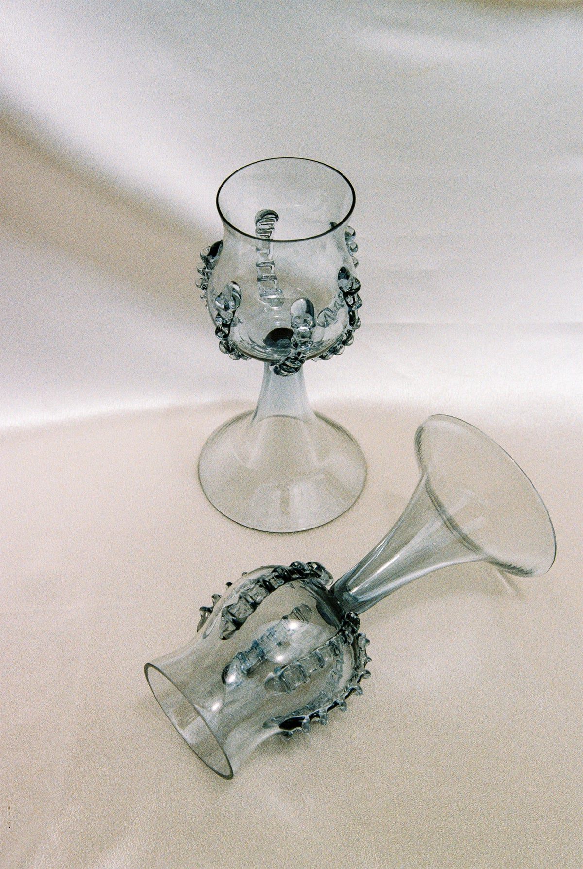 A pair of ribbed, mouthblown Bohemian wine goblets by Lucie Claudia Podrabska—one upright, one on its side—rest atop a smooth, light-colored fabric surface.