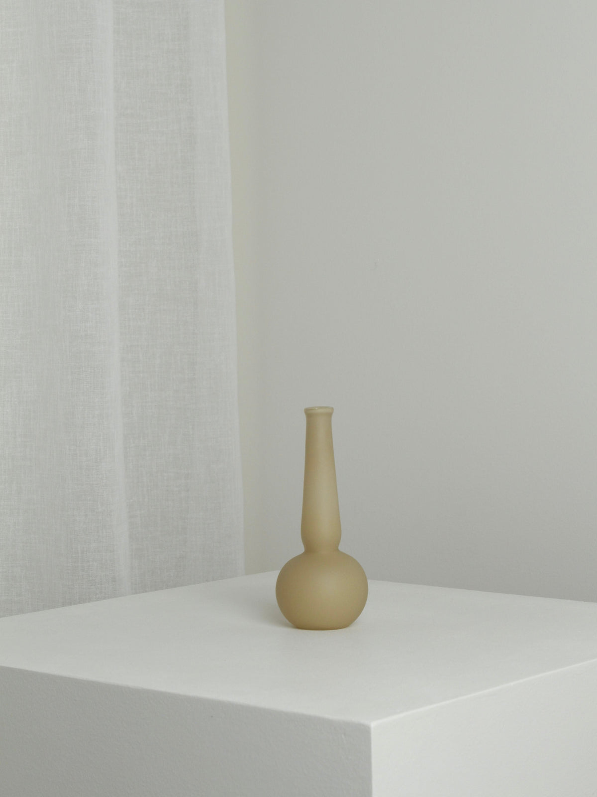 The Verre d'Onge Small Vase in Caramel, featuring a matte finish and long neck, is displayed on a white pedestal against a light wall and sheer white curtain, creating a minimalist, uncluttered setting.
