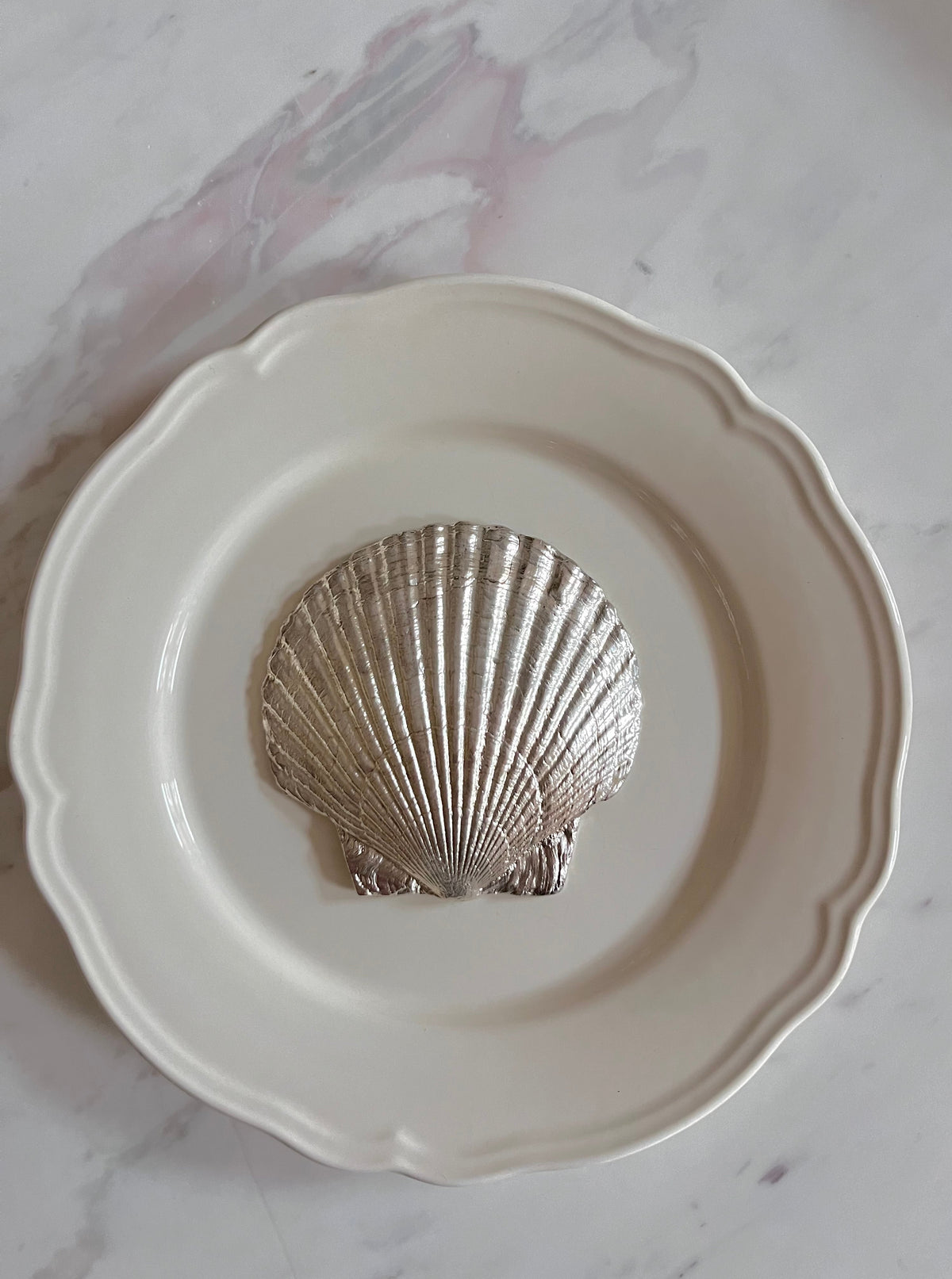 The Médecine Fossil Shell Plate in Silver showcases a metallic silver seashell at its center and a white scalloped edge, perfect as decorative tableware, displayed on a light marble surface.