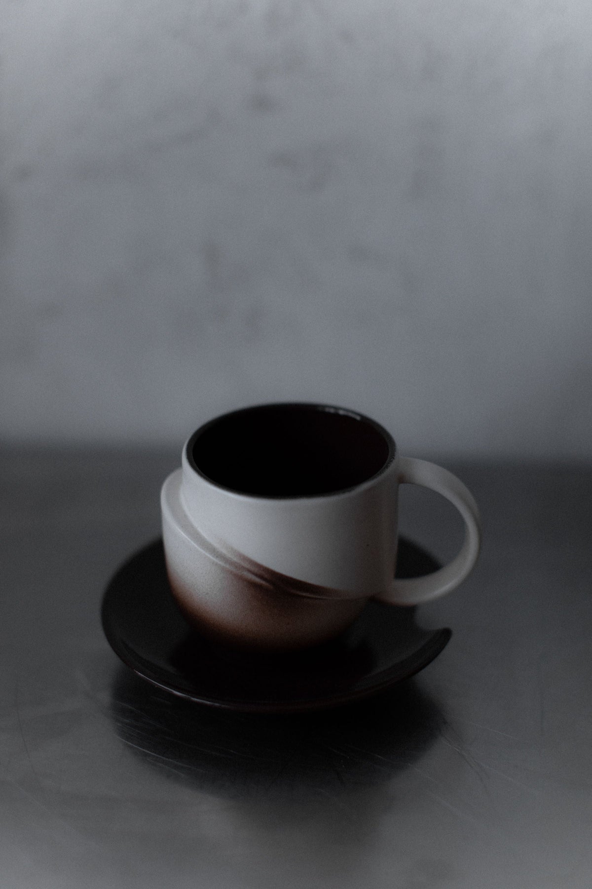 The Art Deco Ceramic set by Out For Lunch features a large-handled vintage coffee cup on a matching saucer in muted brown, resting on a reflective metallic surface with a soft, blurred background.