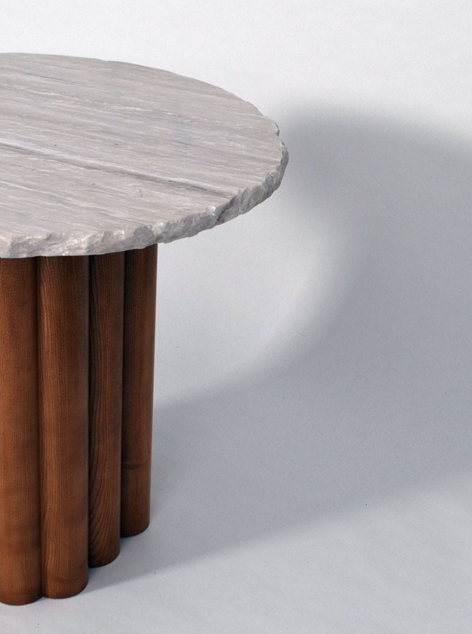 The Carrasqueira Table by Barracuda Interiors features a rough-textured light gray stone top with vertical wooden cylindrical legs, offering an elegant modern look. The image shows part of this unique dining table against a neutral background.