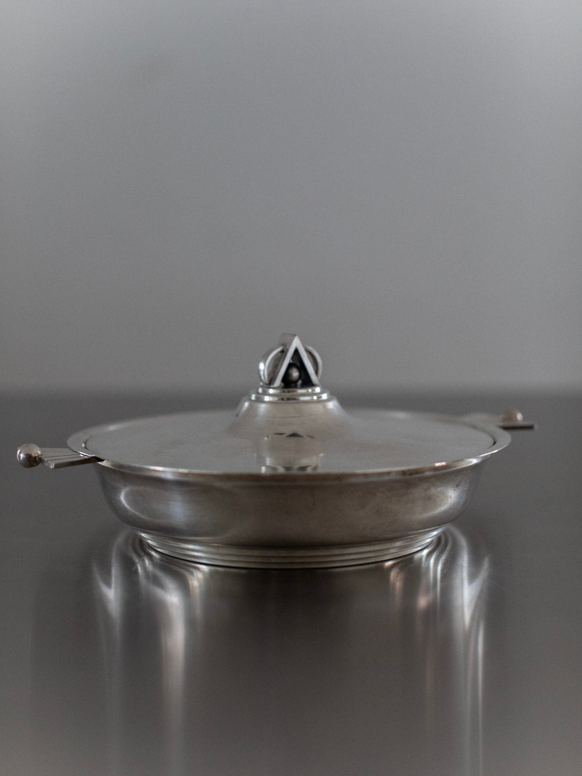 The Out For Lunch Art Deco Bowl, a pre-loved piece with a fitted lid and dual handles, sits on a reflective surface. Its silver finish and triangular handle evoke vintage glamour against the blurred gray background.