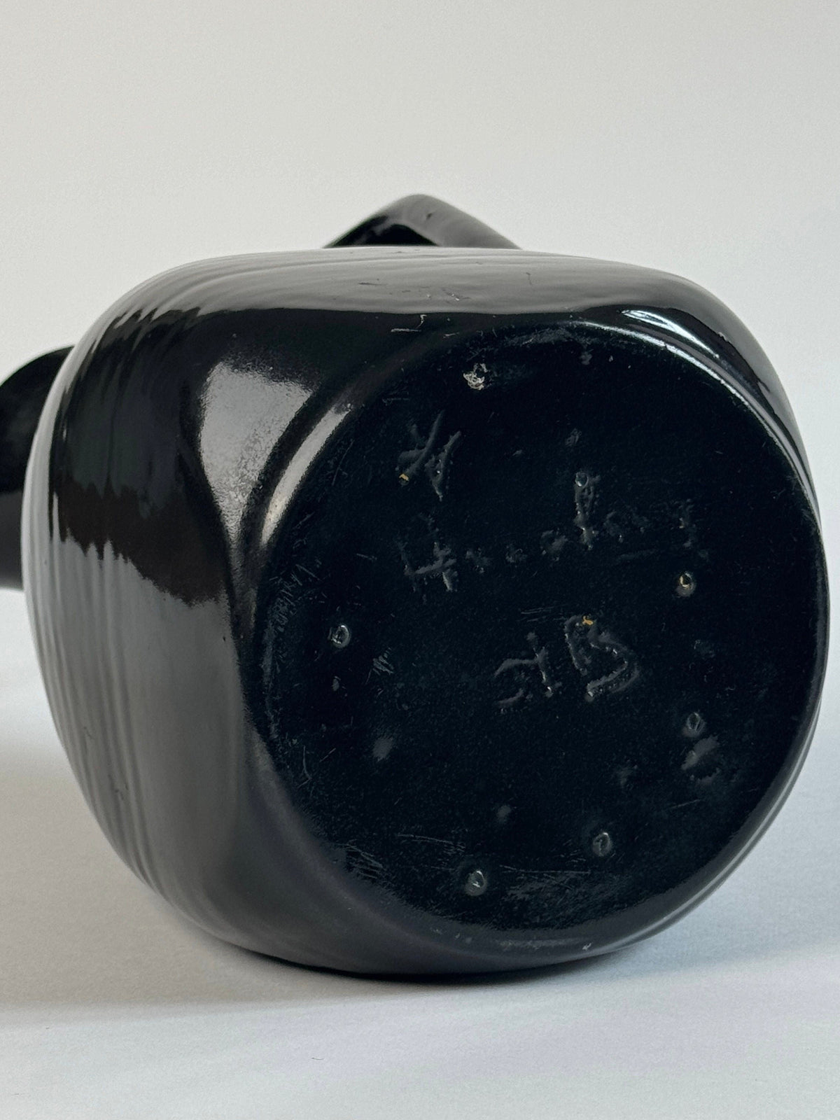 A close-up of the base of a shiny black ceramic piece, likely from the Pair of Accolay Ceramic Carafes by Figuresdesign, showing faint engraved markings and numbers on the bottom surface.