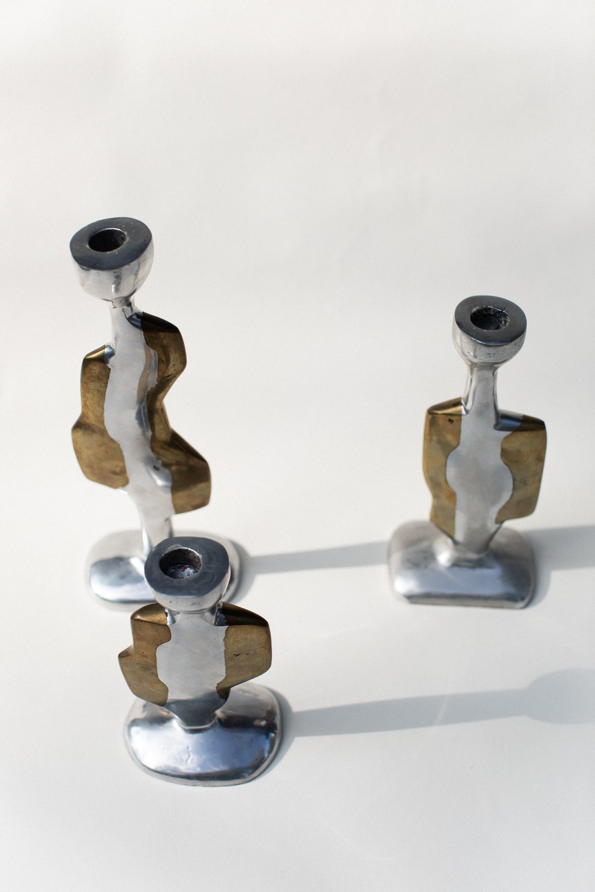 Out For Lunch’s Three Brutalist Candleholders feature abstract, sculptural forms in silver and brass tones, inspired by 70’s brutalist design, beautifully displayed on a light surface with soft shadows.