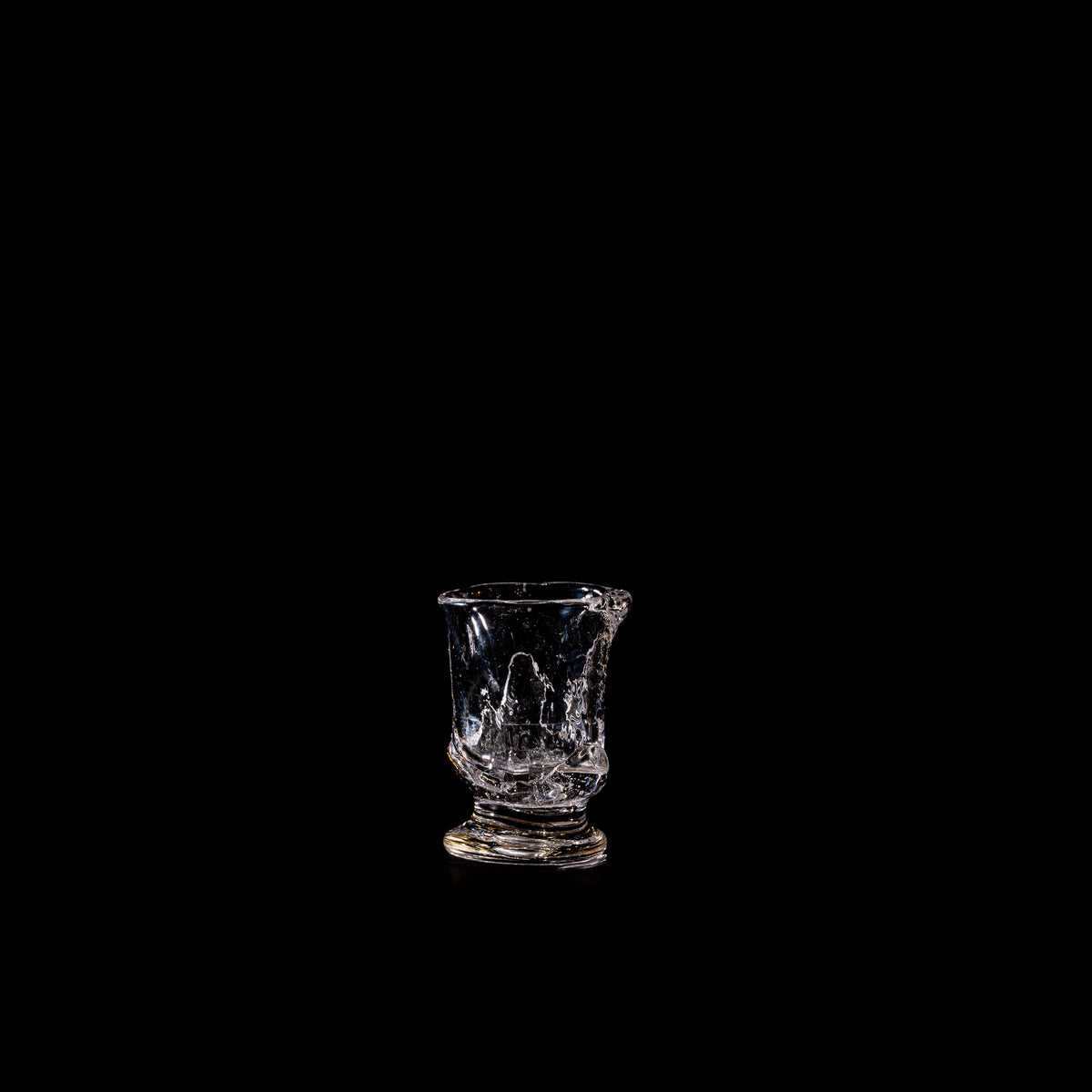 The Organic Shaped Table Glass by Szkło Studio, featuring dramatic cracks and jagged edges, stands alone on a black background under a single light—showcasing the brand’s evocative approach to glassware design.