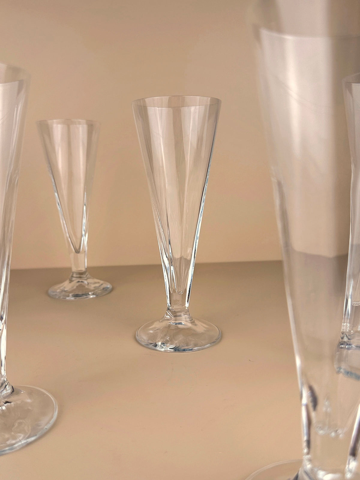 Six empty, clear crystal glasses from the Dodo Vintage Italian Art Deco Revival collection are arranged on a plain, light-colored surface with a beige background, showcasing their elegant shapes and vintage charm.