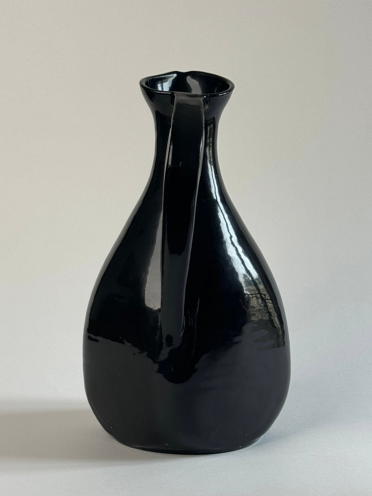 A pair of glossy black Accolay Ceramic Carafes by Figuresdesign, featuring wide bases and narrow necks, shine against a light gray background as their smooth surfaces reflect the light.