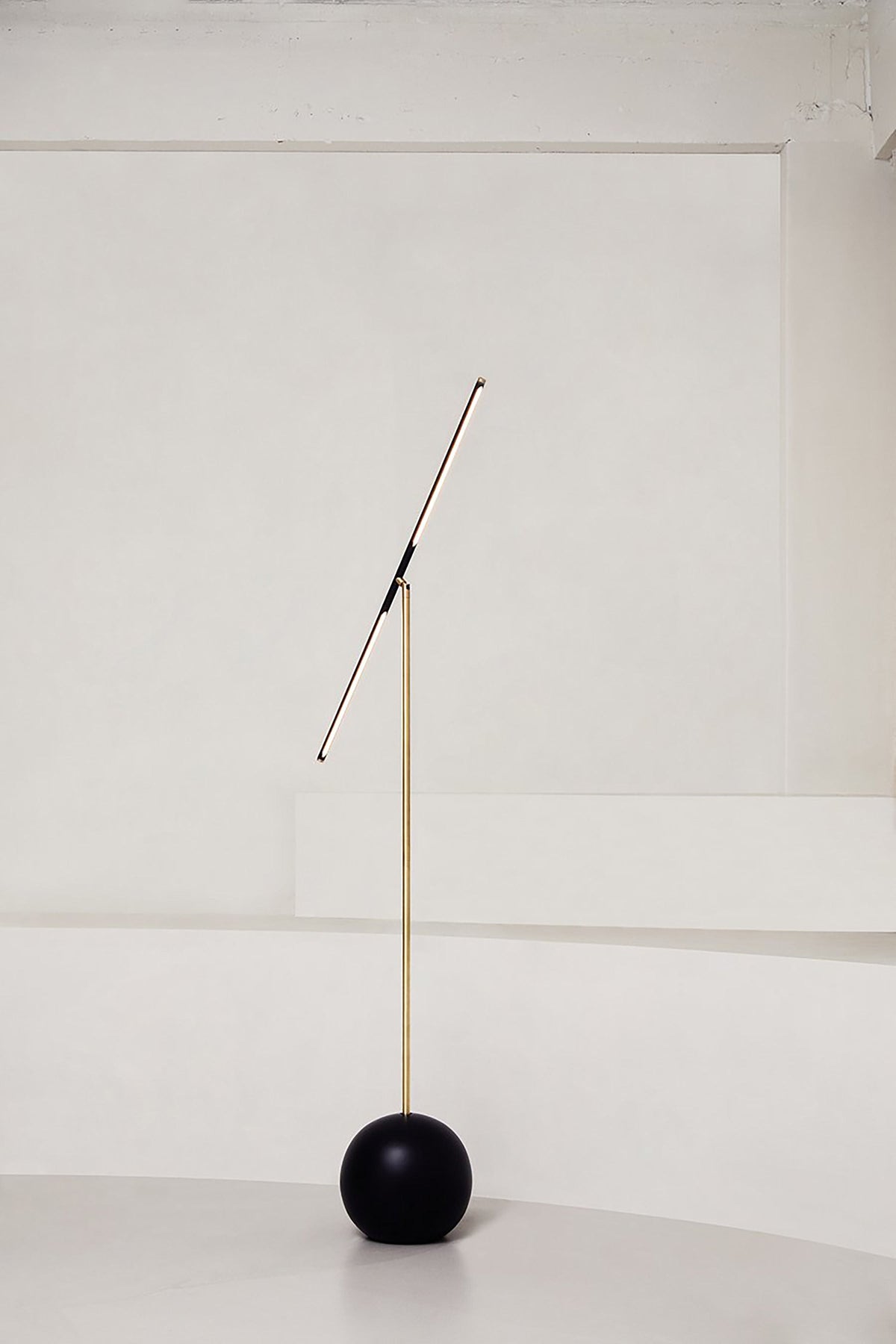 The 'T-Lamp' Floor Lamp by Heako Studio features a minimalist design with a black spherical base, slim gold pole, and angled fixture—an elegant lighting piece shown against a plain white background.