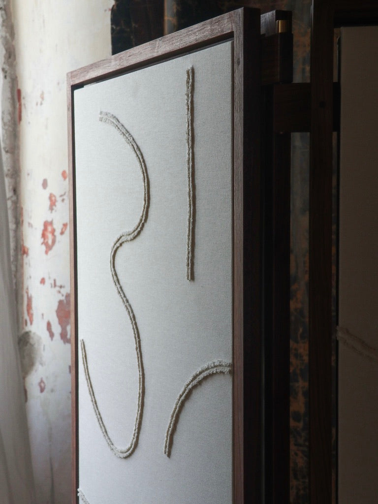 The Cavalletto Room Divider by Filippo Andrighetto features an artistic fabric panel framed in solid walnut, accented with three wavy textured rope lines, set against a rustic wall with peeling paint.