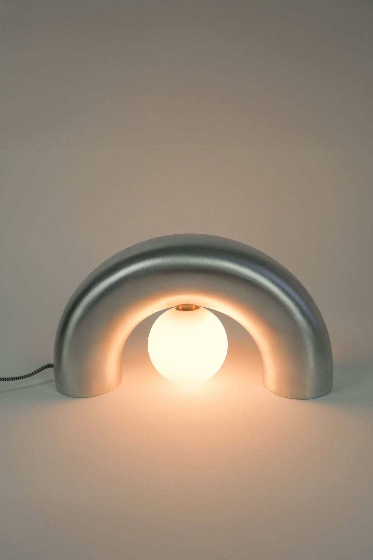 The Arcora Underglow Aluminum Arc Lamp by Heako Studio features a sleek metallic arched base and central glowing bulb, casting soft light for a calming effect against any neutral background.
