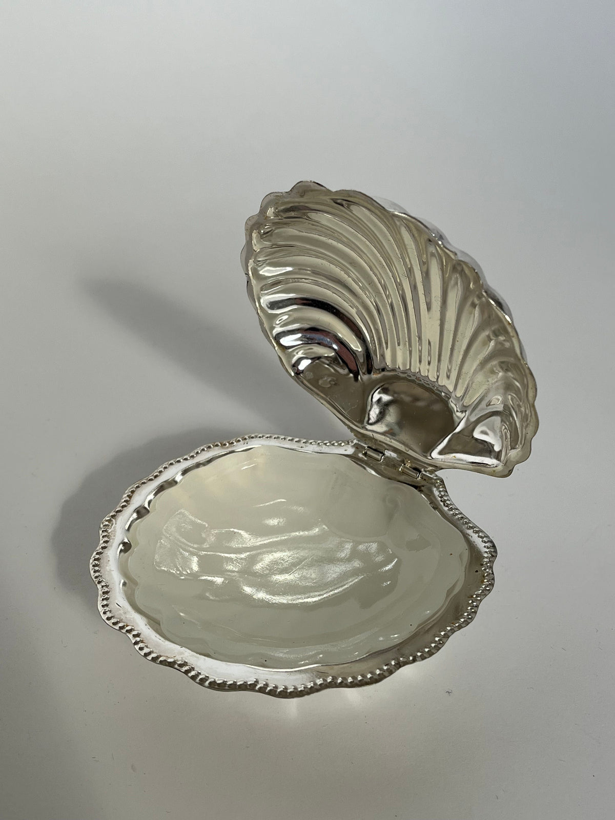 The septembre studios Shell Shaped Butter Dish features a silver, clam shell design with a smooth, reflective interior and scalloped edges, displayed on a light surface.