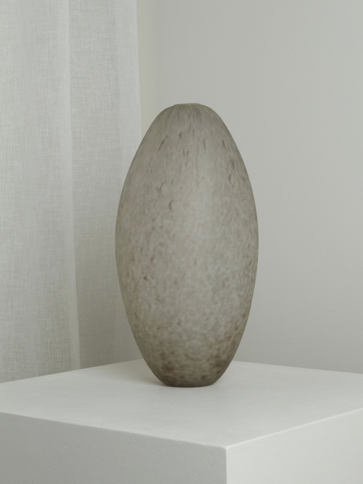 The Verre d'Onge XLarge Vase in Feathered Taupe, with a textured oval shape, sits on a white pedestal against a light gray wall and sheer white curtain.