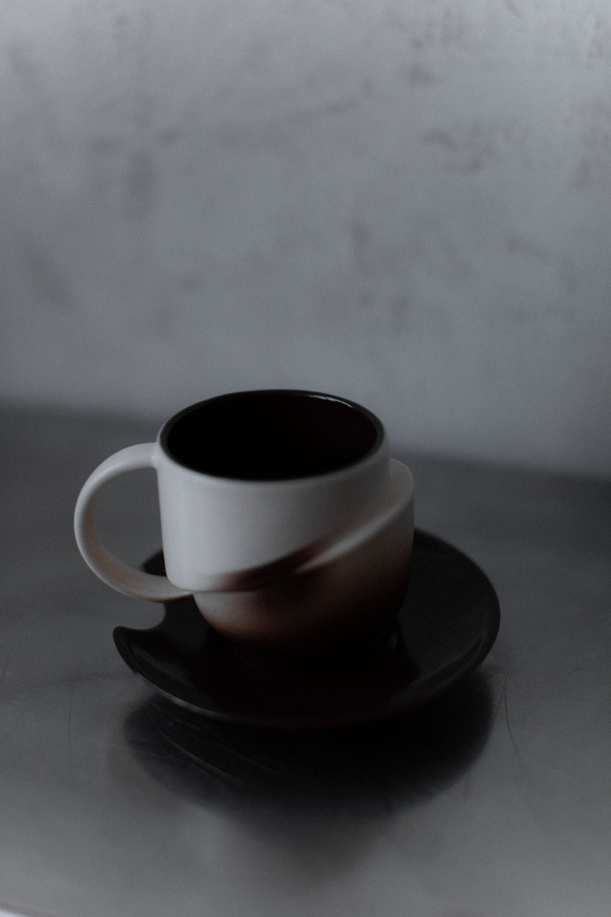 The Out For Lunch Art Deco Ceramic set features a ceramic coffee cup with a unique blurred design, resting on its matching saucer atop a reflective surface against a soft, neutral background.