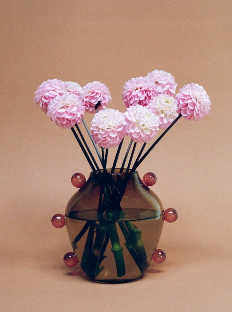 A decorative La Chaleur Vase from Sophie Lou Jacobsen adorned with abstract patterns, containing pink chrysanthemums and ornamented with spherical glass accents on thin stems, set against a pastel peach background.