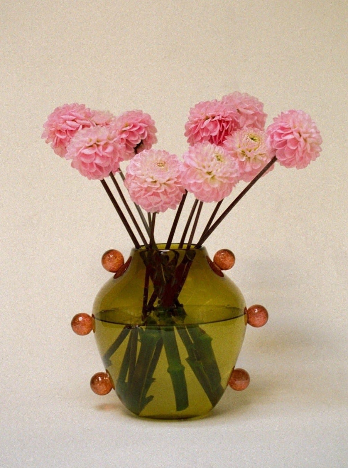 A La Chaleur Vase from Sophie Lou Jacobsen, with a rounded base and a narrow neck, holds a bouquet of pink dahlia flowers. Positioned against a plain background, it creates a serene and minimalist aesthetic ideal for interior decor.