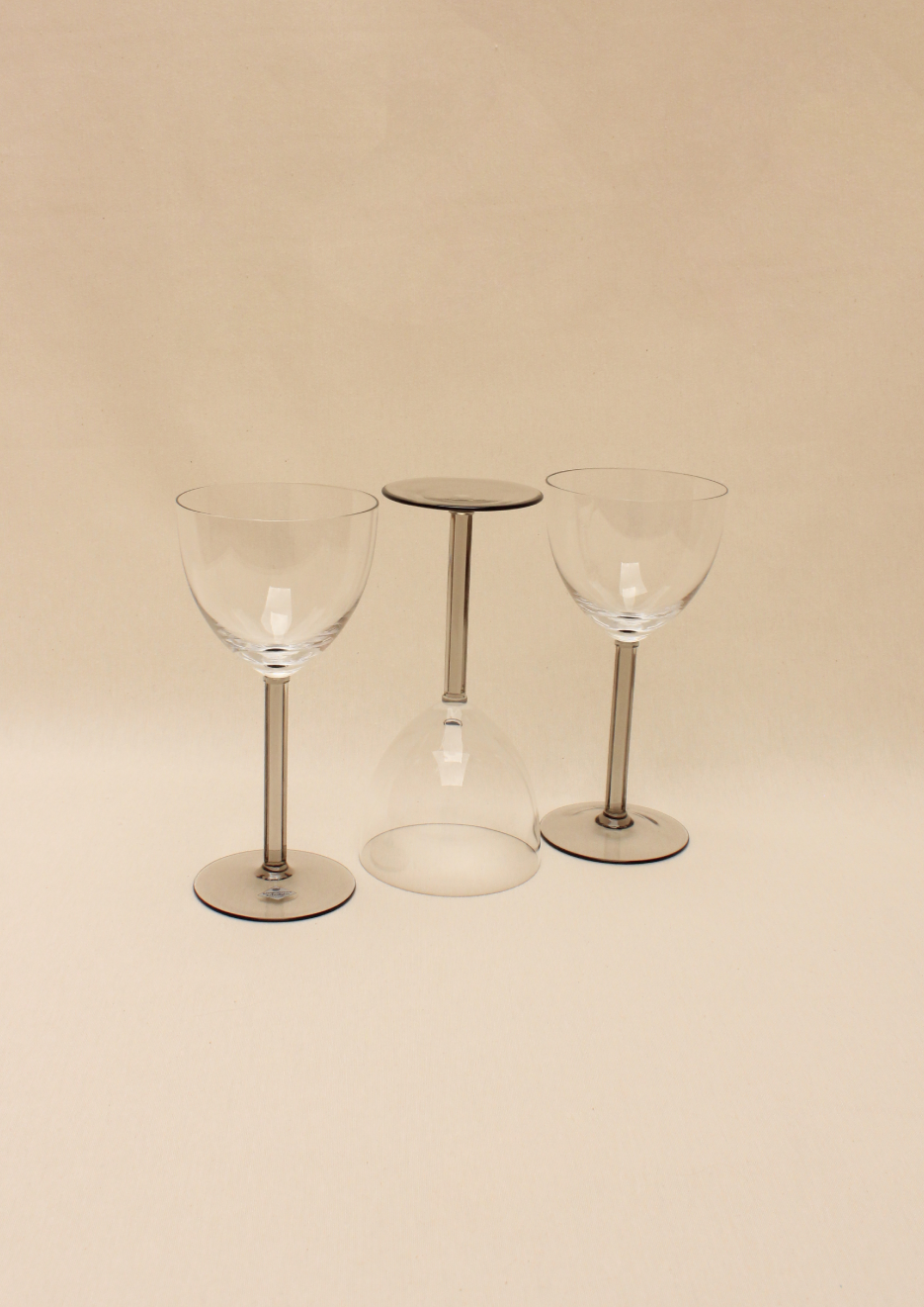 A set of Crystal Wine Glasses from Boga Avante Shop is elegantly displayed on a beige surface, featuring two upright glasses and one upside down in the center. The arrangement highlights a minimalist aesthetic with a luxurious symmetrical touch.