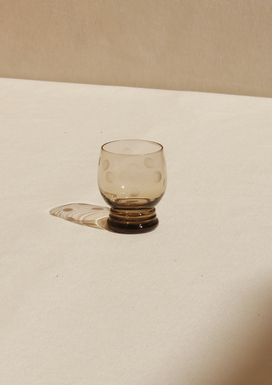 A small, clear vintage Set of Polka Dot Glasses from Boga Avante Shop with abstract patterns placed on a light beige surface, casting a soft shadow in warm, natural light.
