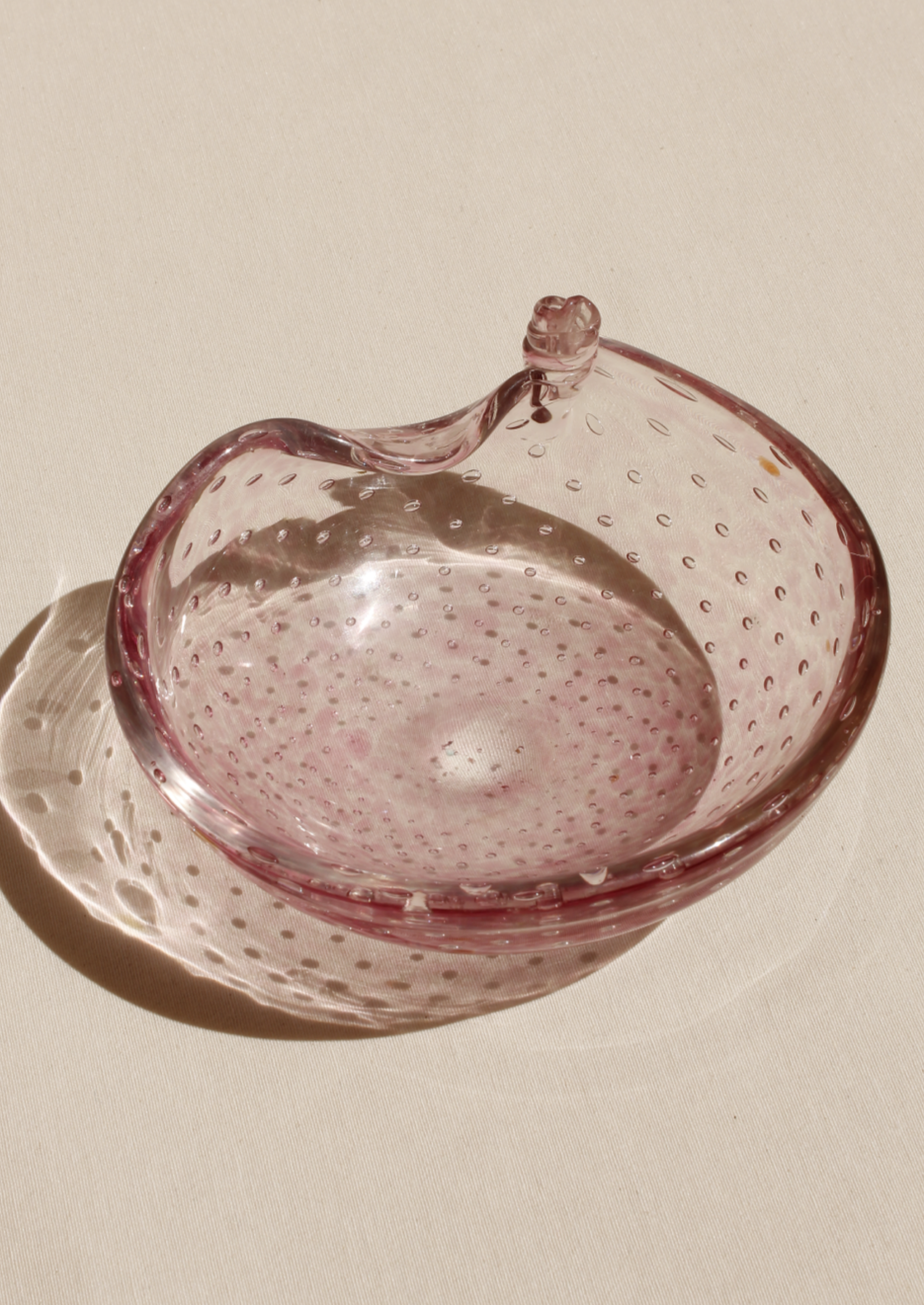 A transparent pink Bullicante Murano bowl shaped like an apple, featuring a textured surface and a small, clear glass stem, placed on a beige fabric background. (Brand name: Boga Avante Shop)