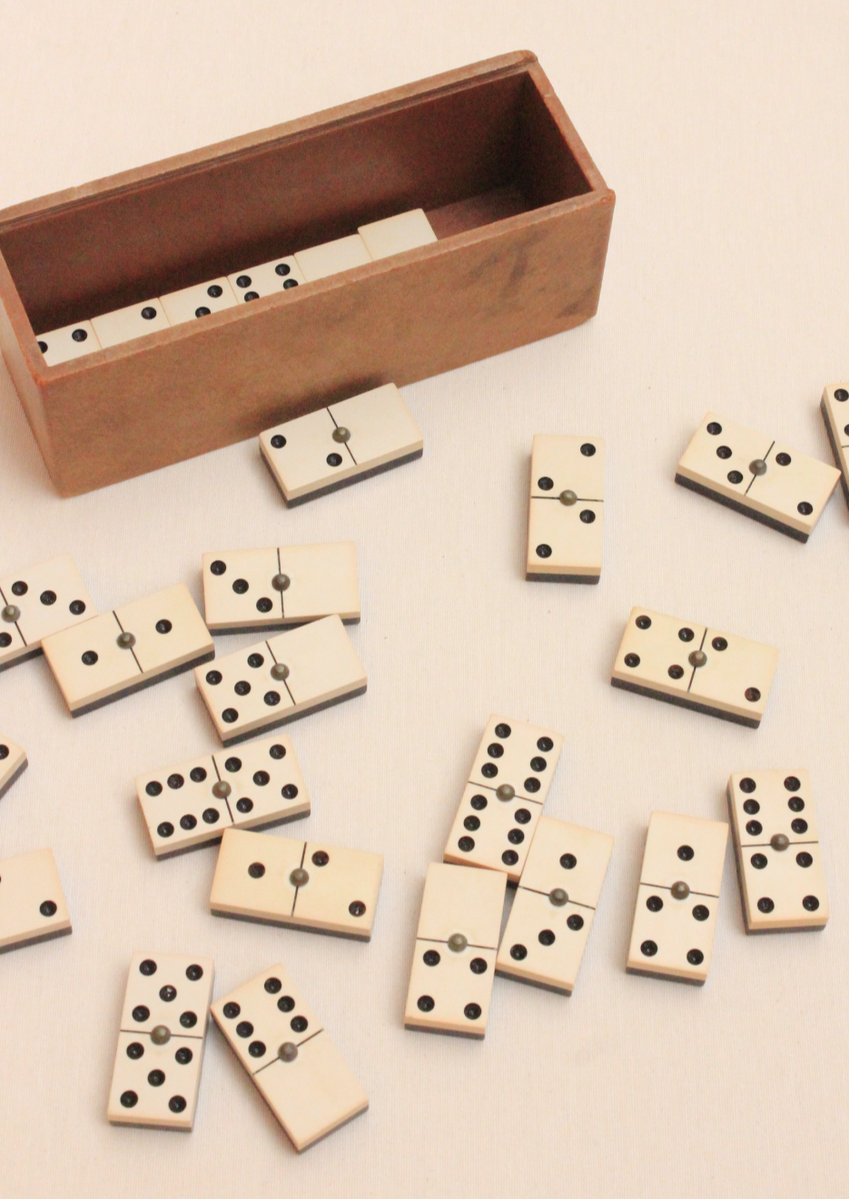 A wooden box with an open lid, spilling out numerous scattered Boga Avante Shop Dominoes onto a cream-colored surface. The dominoes show various numbers of black dots.